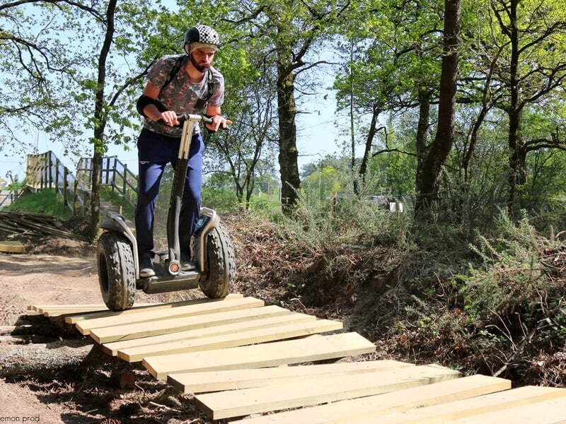 Homme barbu avec casque noir franchissant un pont en bois sur un gyropode dans un environnement forestier.