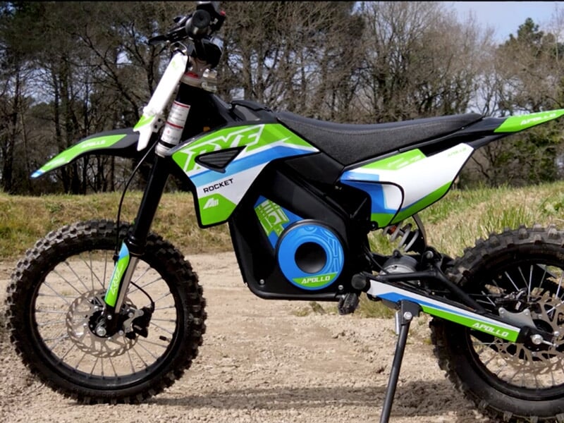 Moto électrique verte, blanche et bleue posée sur un chemin de terre avec des arbres en arrière-plan.