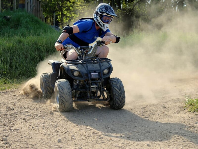Homme en casque et tenue bleue conduisant un quad sur un chemin de terre poussiéreux en forêt.
