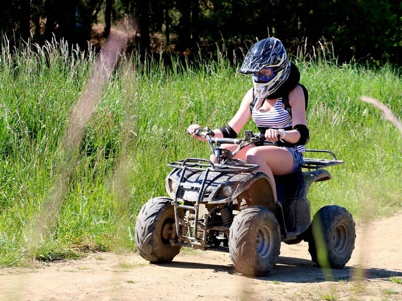 Femme en casque noir et tenue décontractée conduisant un quad sur un chemin de terre entouré d'herbe.