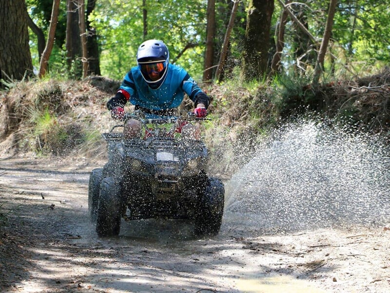 Personne en casque et tenue bleue conduisant un quad qui éclabousse de l'eau sur un chemin boueux en forêt.