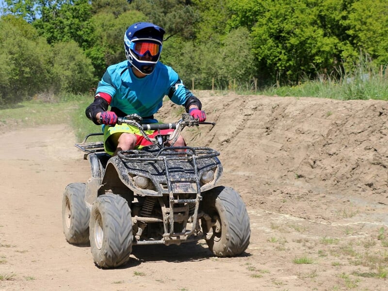 Personne en tenue bleue et casque conduisant un quad sur un chemin de terre dans une zone boisée ensoleillée.