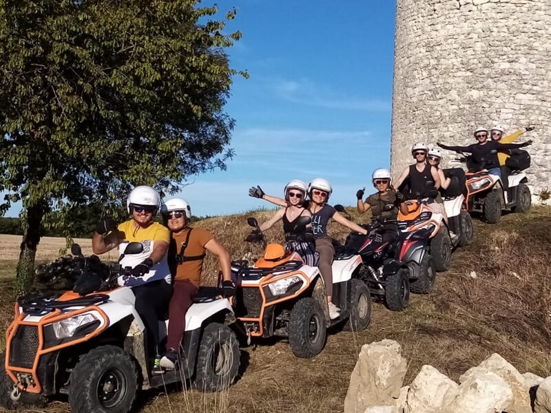 Groupe de personnes souriantes sur des quads alignés près d'une tour en pierre sous un ciel bleu clair.