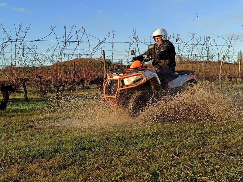 Personne en tenue de protection conduisant un quad dans un champ avec des vignes et éclaboussant de la boue.