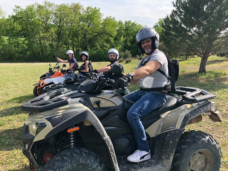 Quatre personnes sur des quads posant dans un champ vert avec des arbres en arrière-plan sous un ciel clair.