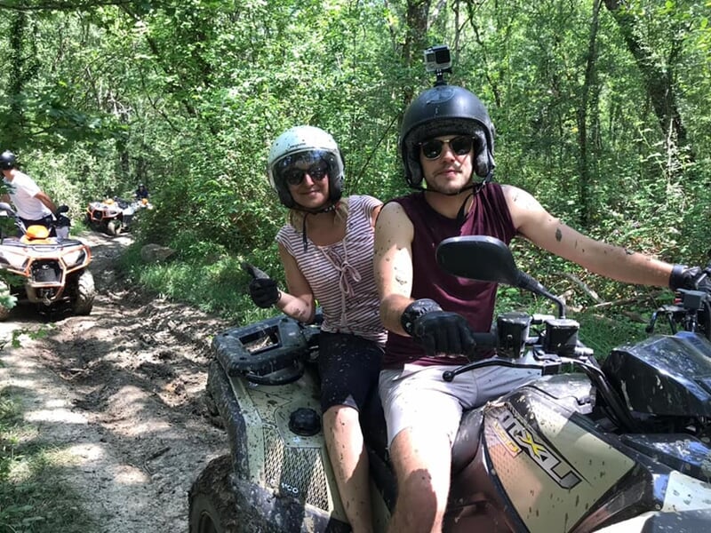 Deux personnes souriantes sur un quad dans un sentier boisé, l'une fait un pouce levé, toutes deux en casque.