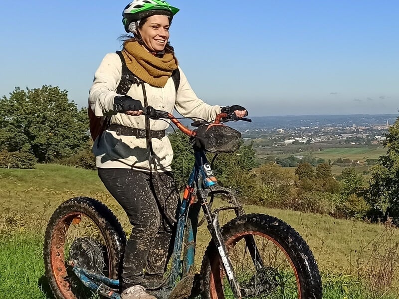 Femme souriante sur un vélo électrique tout-terrain dans un paysage vallonné avec vue sur la ville au loin.