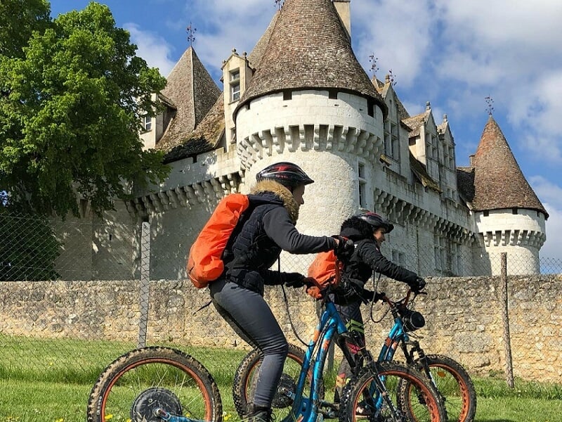 Deux personnes en tenue sportive et casque sur des vélos électriques devant un château médiéval sous un ciel bleu.