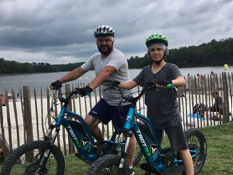 Homme et femme en tenue sportive avec casques posant avec leurs vélos électriques près d'un lac et d'une plage.