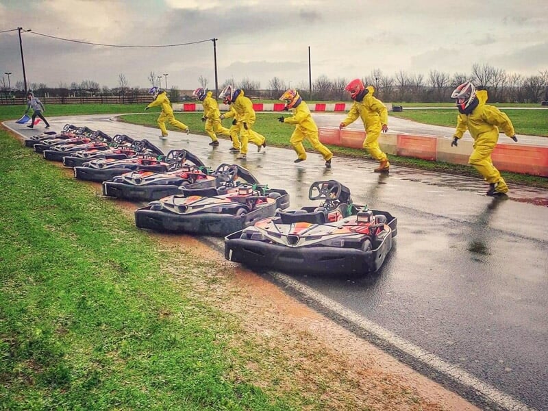 Des pilotes en combinaison jaune sautent pour rejoindre leurs karts alignés sur une piste mouillée de karting.