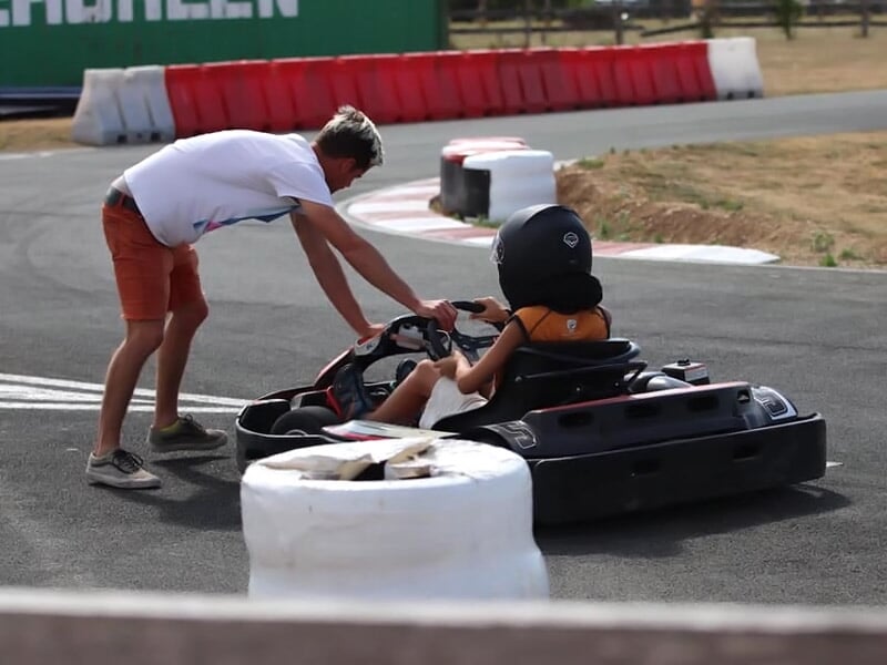 Un instructeur aide un enfant assis dans un kart sur une piste de karting en plein air par temps sec.