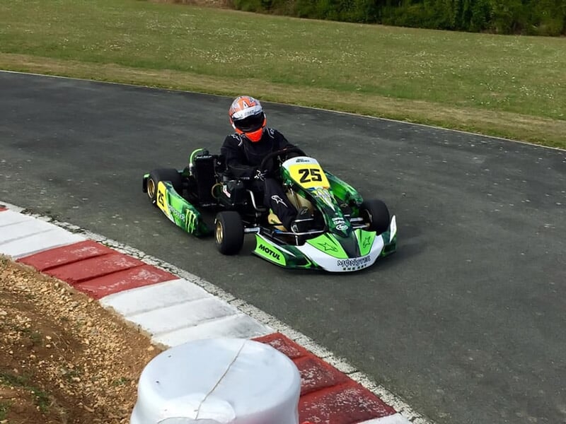 Un pilote en kart vert numéro 25 prend un virage sur une piste de karting entourée d'herbe et de bordures rouges.