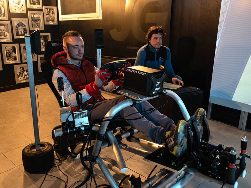 Deux hommes assis dans un simulateur de conduite avec volant, pédales et écran pour entraînement à la conduite.