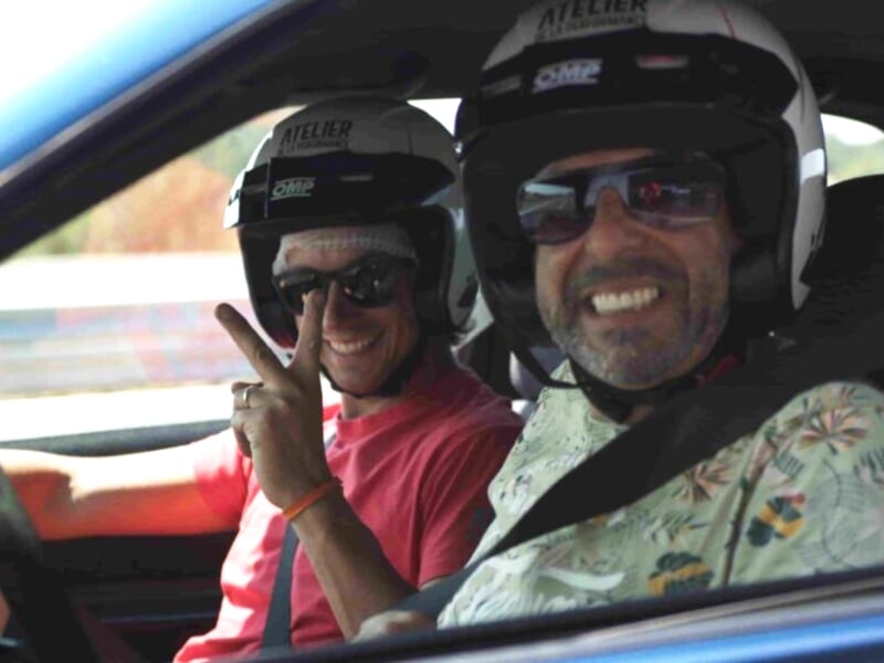 Deux passagers souriants en tenue de pilote avec casques, le copilote faisant le signe de la paix, à l'intérieur d'une voiture de course, ambiance détendue et amicale.