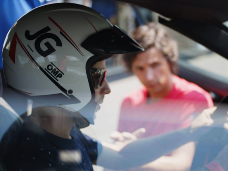 Pilote automobile en casque blanc avec visière abaissée, en discussion à travers la vitre ouverte d'une voiture de sport, autre personne en t-shirt rouge flou en arrière-plan.