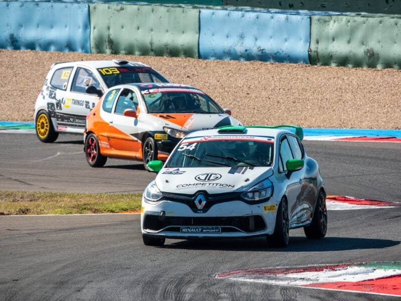 Deux voitures de course, Renault Clio blanche numéro 54 en tête, suivie par une Twingo orange et jaune numéro 103, virage sur piste, autocollants 