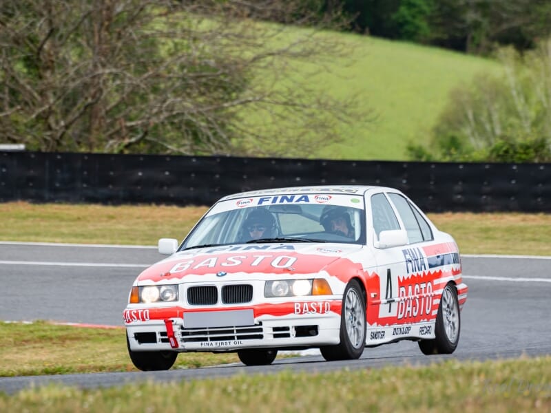 Voiture de course BMW aux couleurs de Basto et Fina, roulant sur une piste avec des arbres et de l'herbe en arrière-plan.