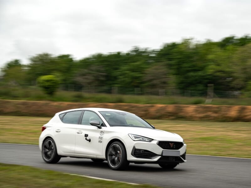 Voiture blanche Cupra sur une piste de course, arbres verts et ciel nuageux en arrière-plan.