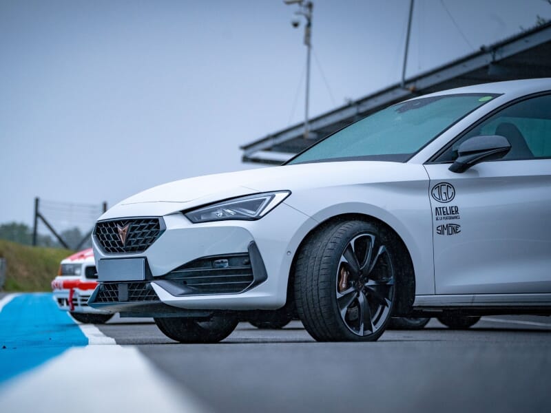 Voiture blanche Cupra garée sur une piste, vue latérale, autre voiture en arrière-plan, ciel nuageux.