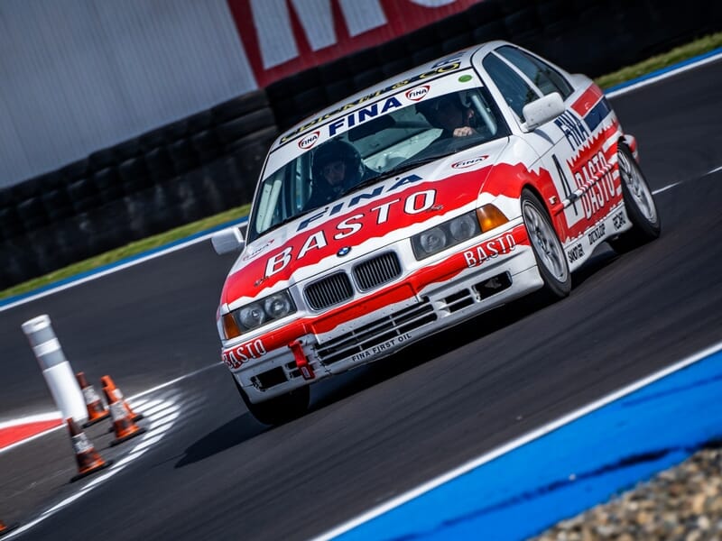 Voiture de course BMW Basto rouge et blanche en virage serré sur circuit avec deux pilotes à bord.