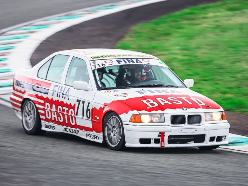 BMW Basto blanche et rouge avec deux passagers en pleine prise de virage sur piste de course asphaltée.
