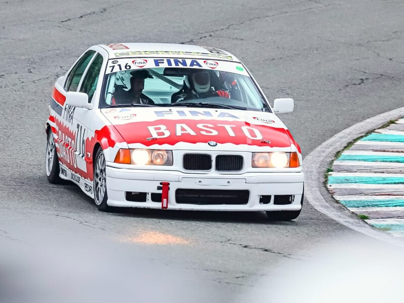 Voiture de course BMW Basto avec deux pilotes, vue de face en virage sur circuit automobile.