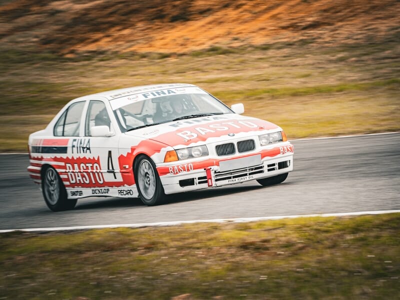 Voiture de course BMW Basto rouge et blanche en virage sur circuit avec paysage naturel en arrière-plan.