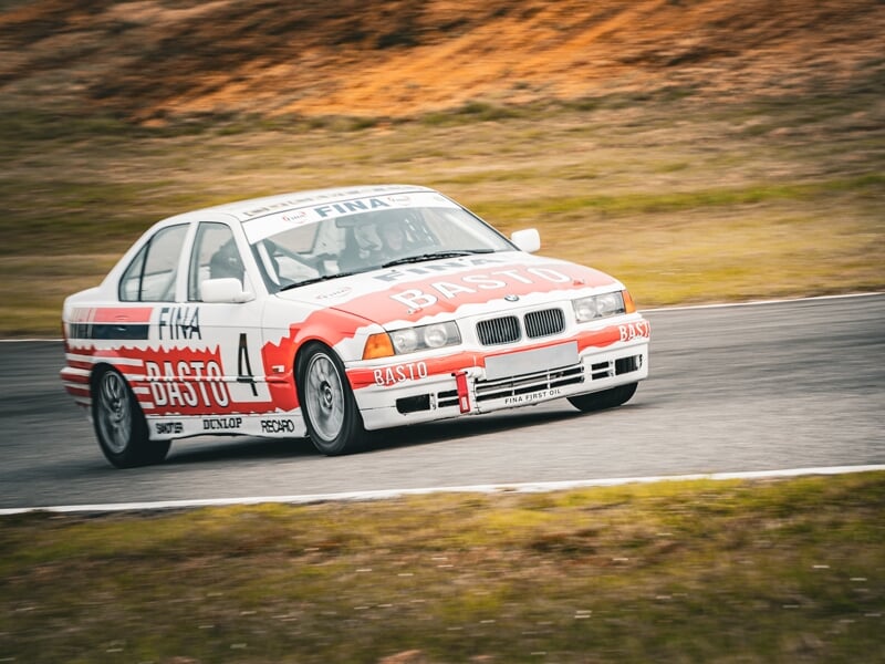 Voiture de course BMW Basto rouge et blanche en virage sur circuit avec paysage naturel en arrière-plan.