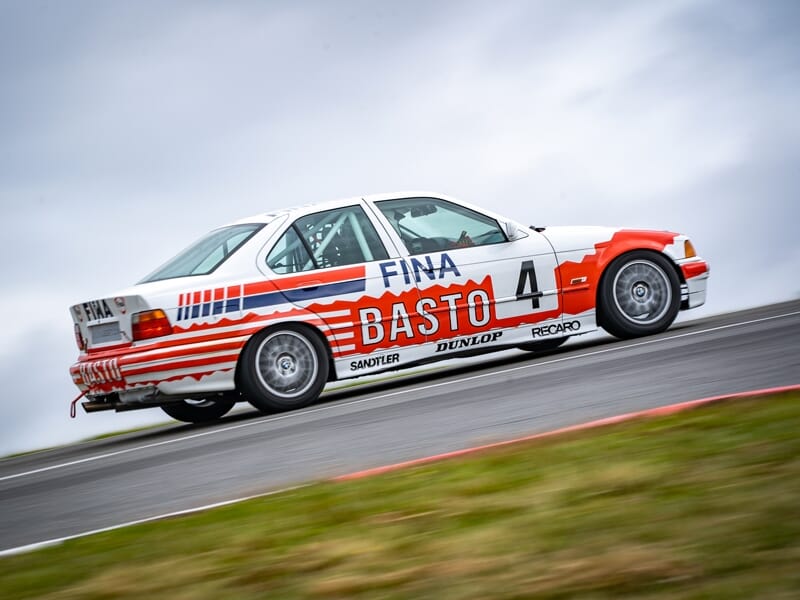 Voiture de course blanche et rouge avec le numéro 4 en pleine accélération sur le circuit de Magny-Cours sous un ciel nuageux.