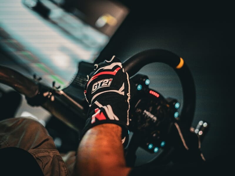 Vue rapprochée d'une main gantée tenant un volant de simulateur de conduite automobile dans un environnement virtuel.