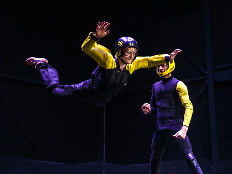Une personne en combinaison de vol jaune avec casque noir exécute un vol contrôlé à l'horizontale face à un instructeur en combinaison bleue et casque jaune, tous deux dans une chambre de vol intérieu