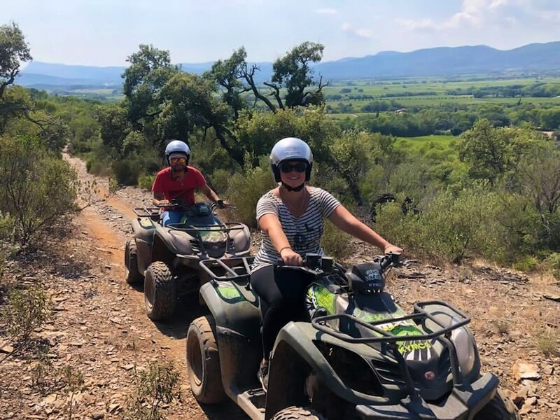 Deux personnes souriantes conduisant des quads sur un chemin rocailleux avec une vue panoramique de la campagne.