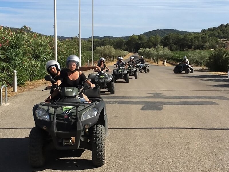 Plusieurs personnes en quad roulant sur une route goudronnée avec des arbres et des collines autour.