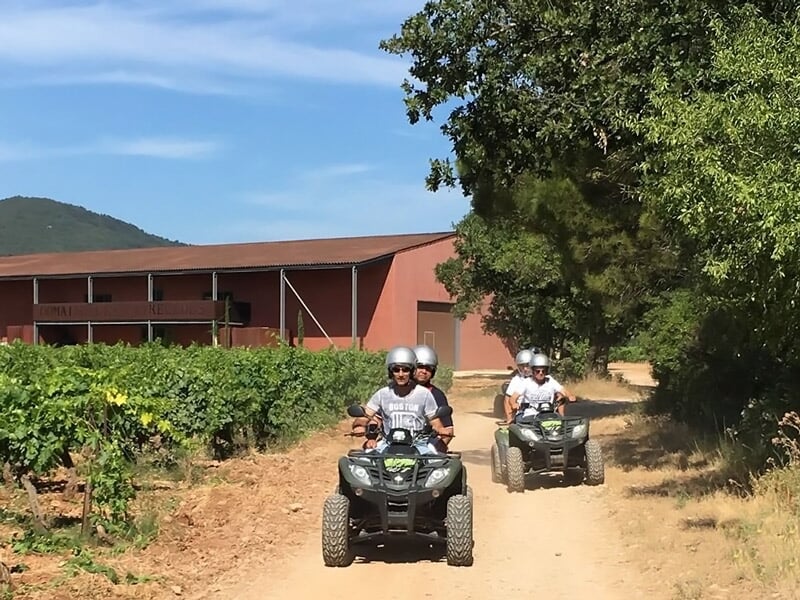 Deux personnes en quad roulant sur un chemin de terre entouré de vignes et d'un bâtiment rouge en arrière-plan.