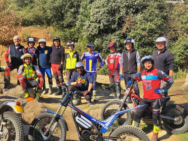 Un groupe de dix personnes en tenue de pilotage et casques posent avec leurs motos électriques sur un chemin en forêt.