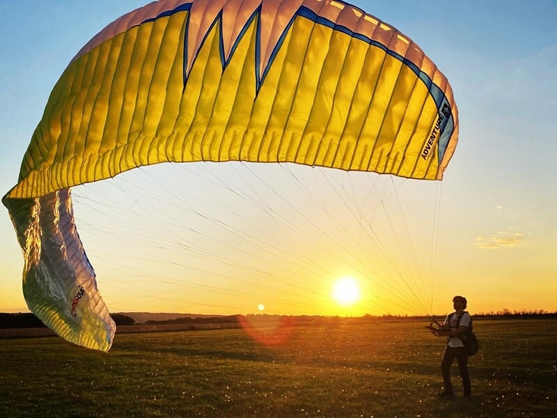 Un homme prépare un parapente jaune au sol au coucher du soleil sur un champ vert.