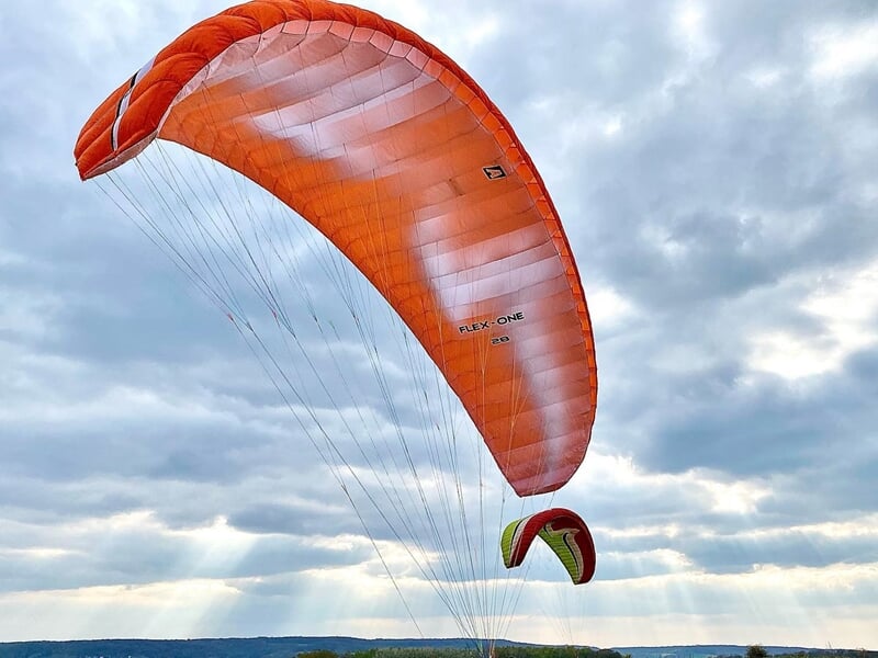 Un parapente orange s'élève dans un ciel nuageux avec un autre parapente visible au loin.