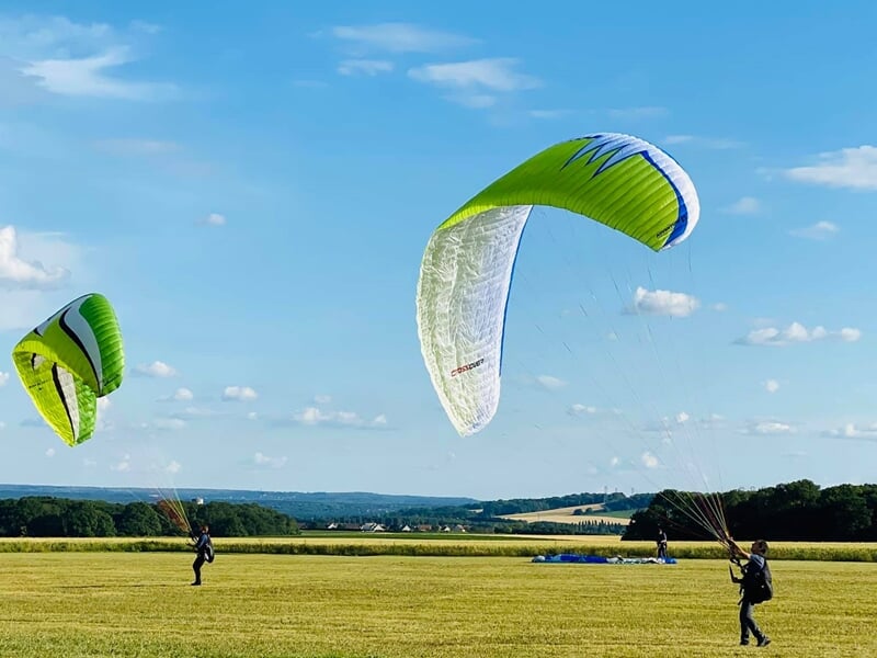 Trois personnes tiennent des parapentes verts et blancs sur un grand champ sous un ciel bleu clair.