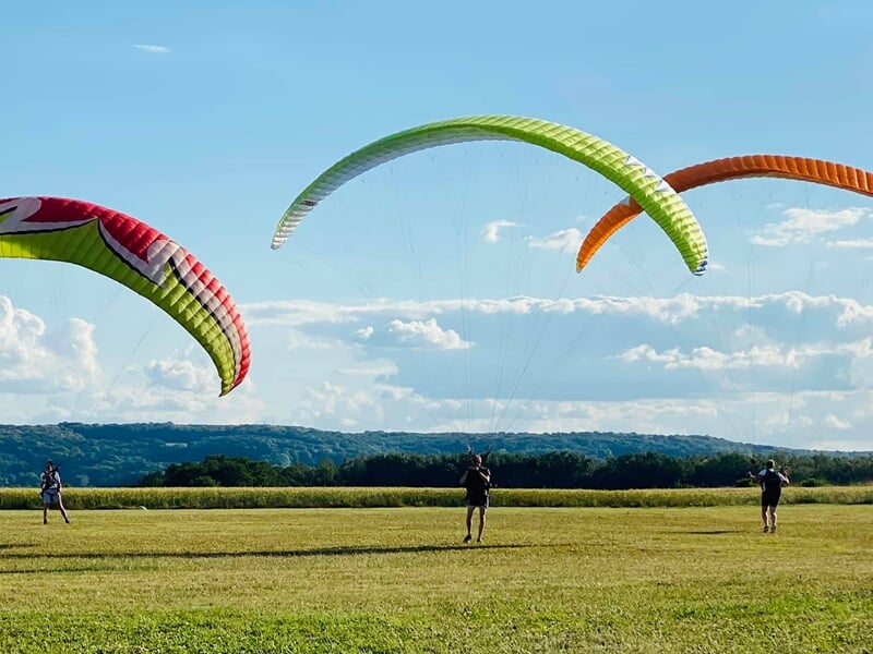 Quatre personnes avec des parapentes colorés s'entraînent sur un champ vert avec un paysage vallonné.
