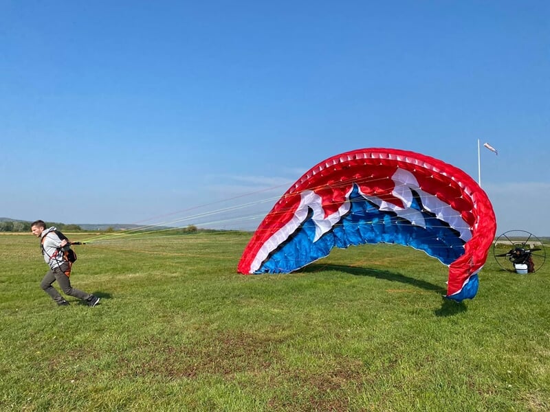 Un homme court sur un champ vert en tenant un parapente rouge, bleu et blanc prêt à décoller.