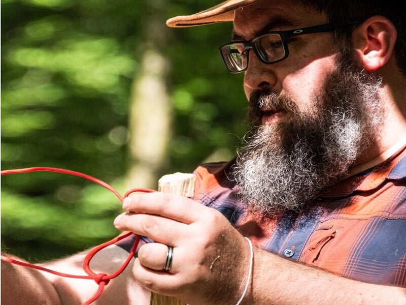 Un homme barbu en chemise à carreaux manipule un outil ou un objet en bois en forêt.