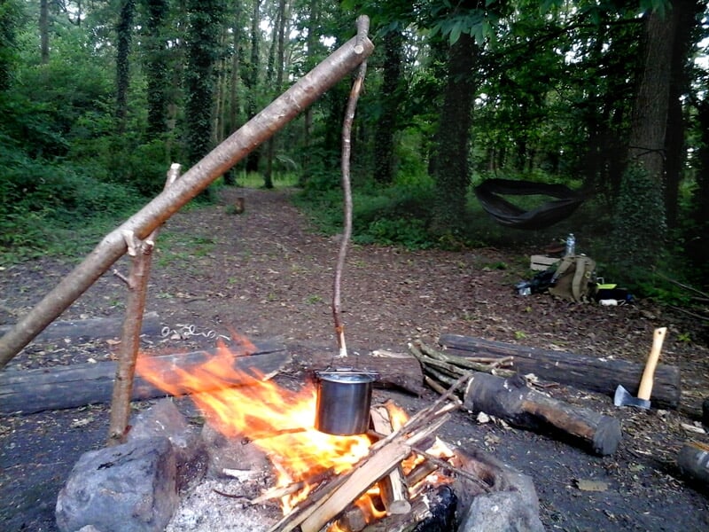 Un feu de camp avec une casserole suspendue au-dessus, entouré de bois et d'outils de camping.
