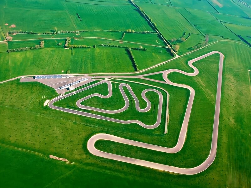 Vue aérienne d'un circuit de moto sinueux entouré de champs verts et d'une zone de paddock avec bâtiments.