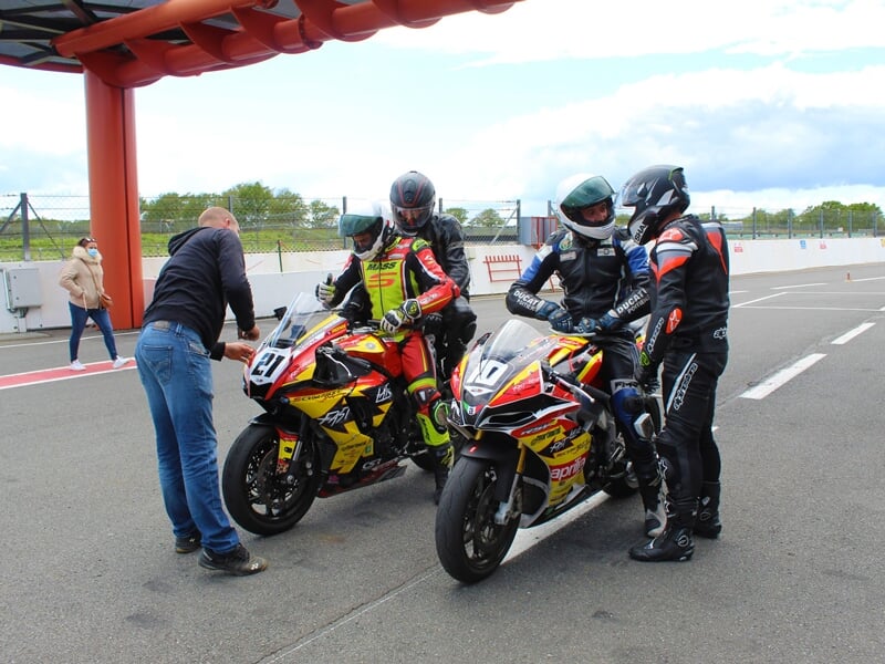 Groupe de motocyclistes discutant sur la piste de course, avec un mécanicien penché sur une moto