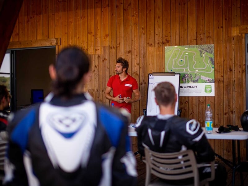 Un moniteur en combinaison de course rouge donnant des instructions devant un tableau avec un circuit de course affiché