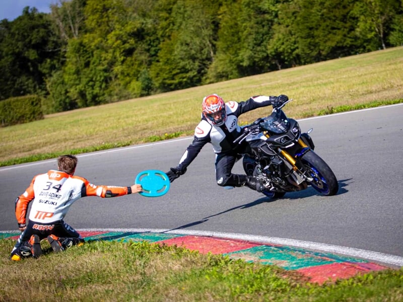 Un pilote en noir et rouge sur une moto sportive noir effectuant un geste de la main à un autre pilote au sol pendant une session de formation sur un circuit
