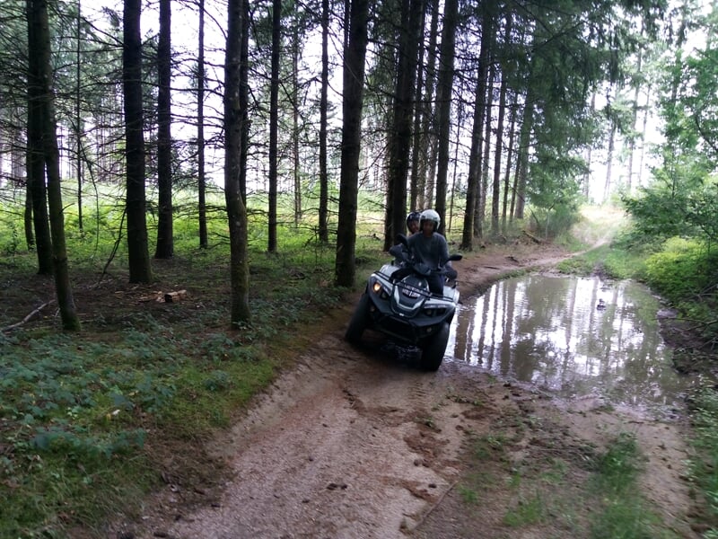 Personne en quad traversant un chemin forestier boueux avec une grande flaque d'eau sur le côté dans une forêt dense.