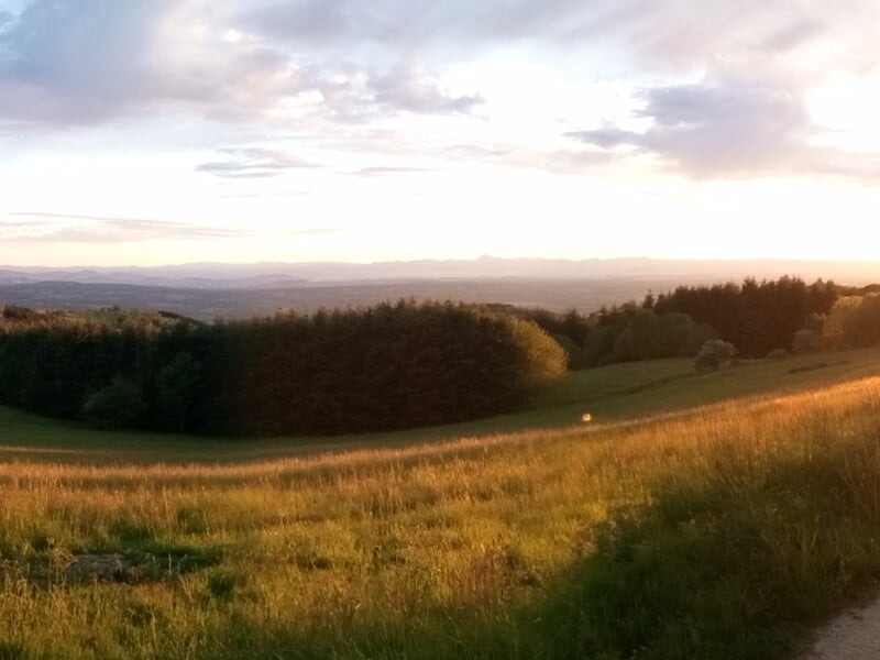 Paysage de campagne au coucher du soleil avec champs verts, collines et forêt à l'horizon sous un ciel nuageux.