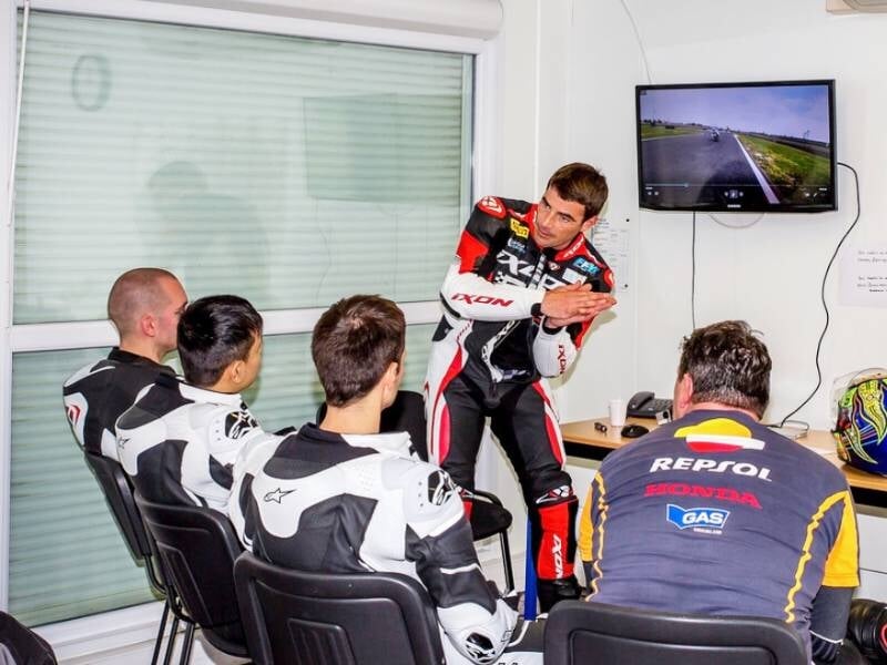 Un instructeur en combinaison de moto explique une technique de pilotage à trois stagiaires assis dans une salle avec un écran.