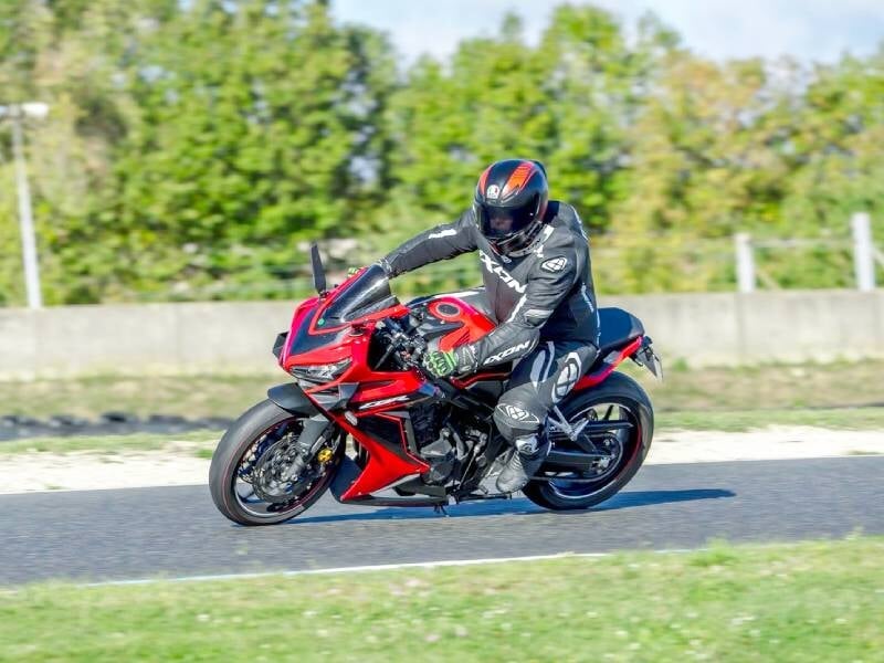 Motard en combinaison noire et casque sur une moto sportive rouge, en virage sur un circuit avec fond d'arbres verts.